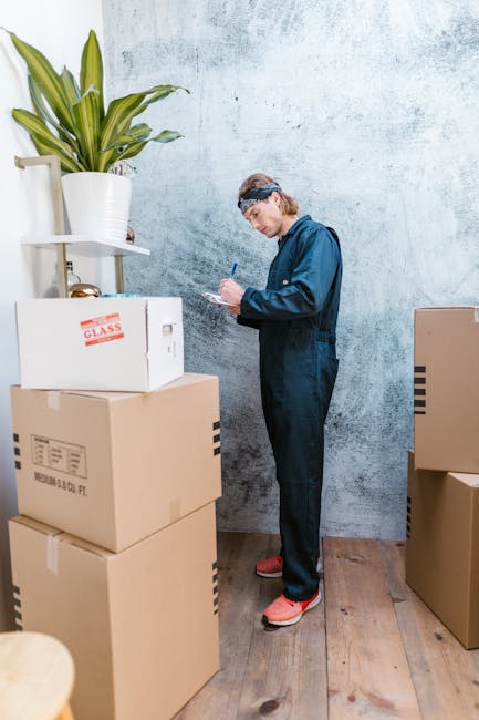 A person dressed in a blue jumpsuit with a headband standing inside a room, surrounded by several stacked cardboard boxes with labels and packaging tape, indicating an ongoing home or office relocatio