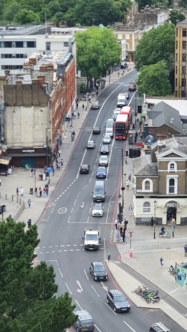An aerial view of a street with a line of various vehicles, including a red double-decker bus, cars, and vans, moving along a curved road in Hampstead. On the sidewalk, pedestrians are walking, some carrying bags or pushing strollers, while others cross at designated crossings. The scene features a mix of residential and commercial buildings, with some structures partially obscured by green trees lining the street. Visible in the foreground are parked bicycles and a small white building with arched windows, indicating urban surroundings. The image captures the busy, traffic-filled environment typical of a home relocation area, where careful furniture transport and moving logistics are essential. Removal Van Hampstead's team may operate in such settings to facilitate house removals with small or tight access routes, utilizing appropriate loading and packing procedures to ensure a smooth moving process.