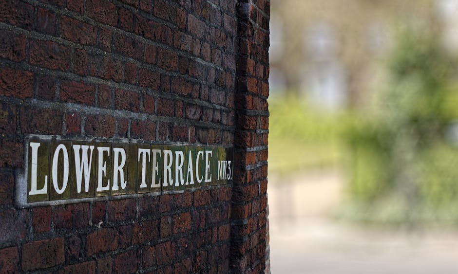 A close-up view of a brick wall with a street sign affixed to it, reading 'Lower Terrace NW3' in white letters on a green background. The brickwork is dark red with visible mortar joints, and the sign appears slightly weathered. In the background, part of a narrow, paved pathway is visible, leading past greenery and blurred trees, indicating an outdoor residential area. Inside a property, situated nearby the wall, are several cardboard boxes, a wooden piece of furniture, and a plastic-wrapped appliance, all set on a wooden floor. Two individuals, possibly removal staff, are engaged in a home relocation process, lifting and maneuvering a large piece of furniture, utilizing a dolly and straps. A spacious van with an open rear door is partially visible on the pavement, ready for the furniture transport, with protective blankets covering items. The scene captures a house removal operation with a focus on detailed packing, loading, and transport activities within a tight-access property, as managed by Removal Van Hampstead.