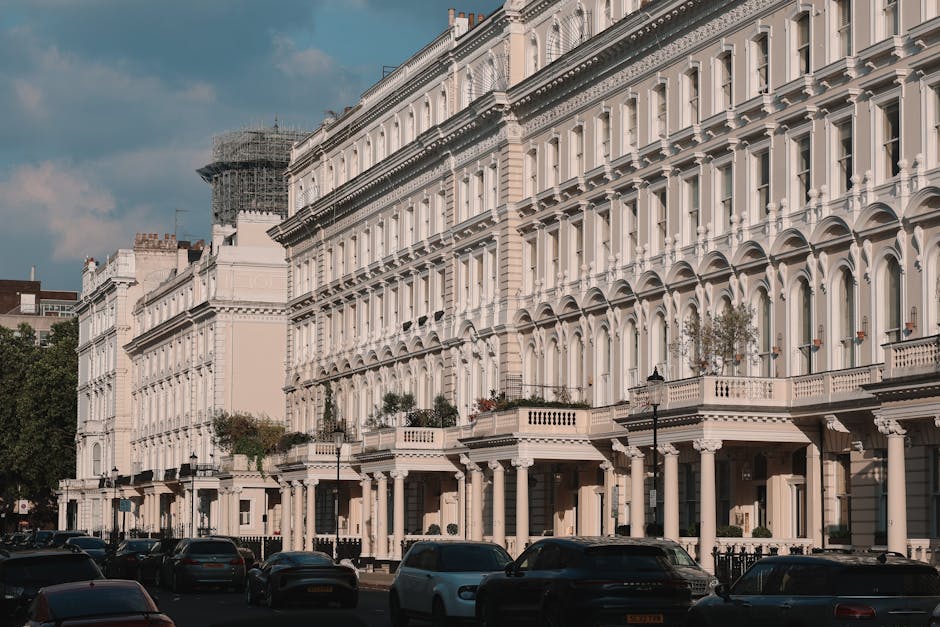A row of white Victorian terraced houses with ornate architectural detailing, including arched window frames and decorative cornices, situated along a street with parked cars in front. The buildings feature balconies with balustrades and potted plants on some ledges. A black lamppost is visible on the pavement, and the scene is illuminated by natural daylight with a partly cloudy sky overhead. In the background, scaffolding surrounds a building under renovation. The street scene captures the typical residential environment where house removals and furniture transport are undertaken, and [COMPANY_NAME] specializes in home relocation and packing services, often involving careful loading of furniture and boxes onto vans positioned at the curb for efficient moving logistics.
