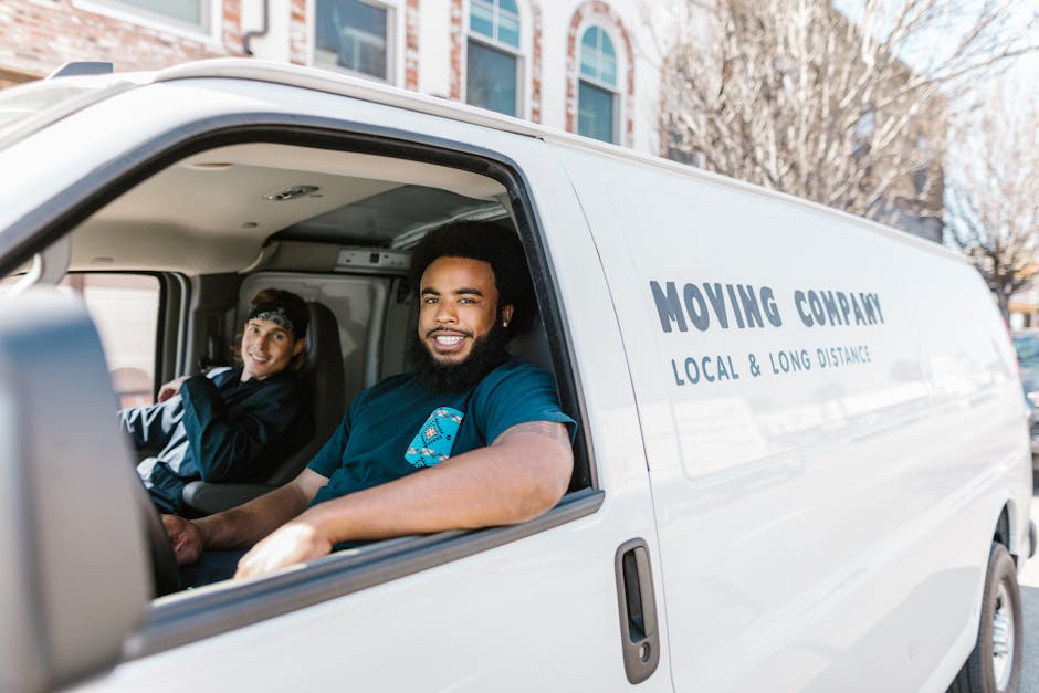 Inside a white moving van labeled 'Moving Company Local & Long Distance', two movers are seated in the cab, ready for home relocation tasks. The driver, a man with dark curly hair and a beard, is smiling and wearing a navy blue T-shirt, while the passenger, a woman with dark hair in a headband, is also smiling. Visible through the open window are cardboard boxes and wrapped furniture pieces stacked inside the van, indicating packing and furniture transport for a house move. The van is parked on a residential street with modern multi-story buildings and leafless trees in the background, suggesting a typical urban move in Hampstead. Equipment such as straps or moving blankets might be present, although not clearly visible, and the interior of the van shows essential packing materials and tools used during the loading process. The scene demonstrates the professionalism and readiness of Removal Van Hampstead for efficient house removals, highlighting the logistics involved in a Victorian terrace move, including safe loading, packing, and transportation.