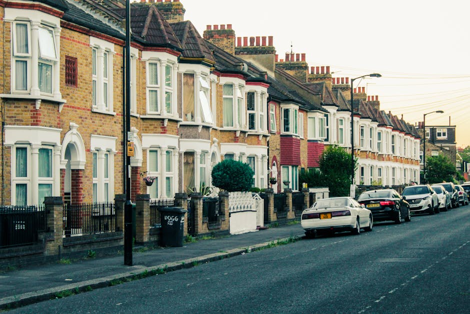 A row of white Victorian terraced houses with ornate architectural detailing, including arched window frames and decorative cornices, situated along a street with parked cars in front. The buildings feature balconies with balustrades and potted plants on some ledges. A black lamppost is visible on the pavement, and the scene is illuminated by natural daylight with a partly cloudy sky overhead. In the background, scaffolding surrounds a building under renovation. The street scene captures the typical residential environment where house removals and furniture transport are undertaken, and [COMPANY_NAME] specializes in home relocation and packing services, often involving careful loading of furniture and boxes onto vans positioned at the curb for efficient moving logistics.